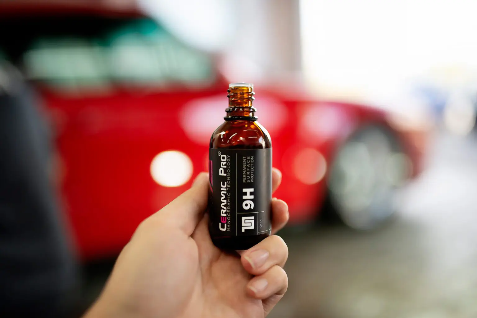 Hand holding Ceramic Pro bottle, red car background.